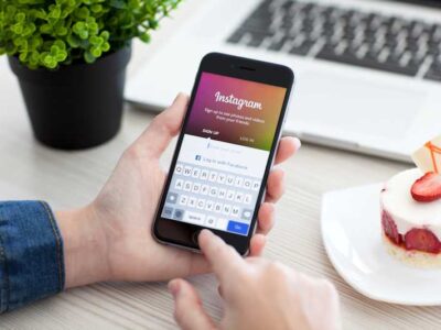 Hands holding mobile device with Instagram logo next to a laptop and strawberry cheesecake