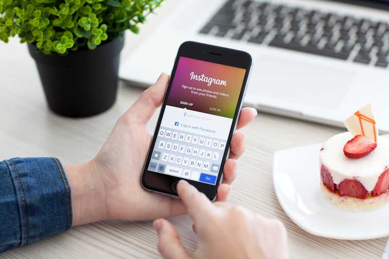 Hands holding mobile device with Instagram logo next to a laptop and strawberry cheesecake